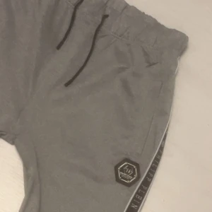 Grå mjukisbyxor från Philipp Plein - Grå mjukisbyxor från Philipp Plein med svarta dragsnören i midjan och elastiska benslut. Snygg logga på benet och svart sidoband med märkets namn längs hela benet. Perfekta för en chill och sportig look.