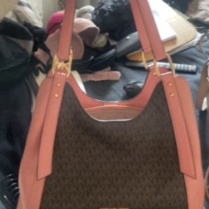 Michael Kors axelväska med rosa detaljer - Snygg axelväska från Michael Kors med klassiskt MK-mönster i mörkbrunt och svarta toner. Väskan har breda axelband i rosa skinn och guldfärgade metalldetaljer. Perfekt storlek för att få plats med allt du behöver och ger en lyxig känsla till din outfit. Kommer inte till användning, prislapp kvar! Nypris 6000kr. Säljer för 2000kr