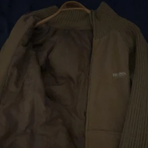 Brun stickad jacka från HUGO BOSS - Säljer en brun jacka från BOSS med stickade ärmar och krage. Jackan har en enkel design med diskret BOSS-logga på bröstet och stora fickor framtill. Perfekt för dig som gillar stilren och bekväm look. Dock är dragkedjan trasig 