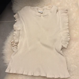 Beige ribbad topp med volang från Zara - Supersöt ribbad topp från Zara i beige med volangdetaljer längs ärmhålen och nederkanten. Toppen är ärmlös och har en mjuk, stretchig känsla som gör den riktigt skön att bära. Perfekt för dig som gillar en stilren men ändå lekfull look.