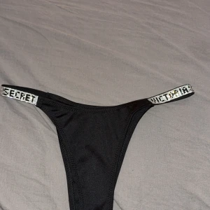 Svart bikini från Victoria's Secret - Snygg svart bikini från Victoria's Secret med glittriga axelband där märkesnamnet står i stenar. Klassisk modell med smala band och lyxig känsla, perfekt för stranden eller poolen. Materialet är mjukt och följsamt för bästa komfort.