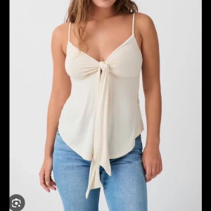 Beige linnetopp med knyt från Gina Tricot - Supersöt beige linnetopp från Gina Tricot med smala axelband och snygg knytning framtill. Toppen har en lätt v-ringad urringning och är tillverkad i mjuk bomull, perfekt för varma dagar. Snygg att styla med jeans eller kjol!