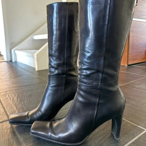 Svarta höga skinnstövlar med klack - Säljer ett par svarta Italienska stövlar i äkta skinn med hög klack och fyrkantig tå. Stövlarna har en stilren och klassisk look, perfekt till jeans eller kjol. De är höga och har en elegant siluett som ger en cool vibe. Sparsamt använda.