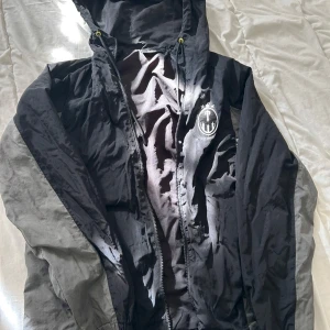 Svart och grå vindjacka med huva - Svart och grå vindjacka med huva och dragkedja framtill. Jackan har ett vitt emblem på bröstet och gula detaljer vid snörningen. Tillverkad i lätt och glansig polyester, perfekt för blåsiga dagar. Snygg och sportig look med normal passform.