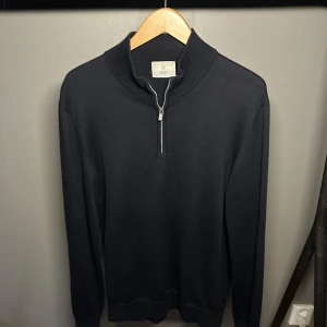 Mörkblå stickad tröja med half zip - Svart stickad tröja från Bläck med hög krage och snygg half zip-dragkedja framtill. Tröjan har långärmad passform och ribbade muddar vid ärmslut och nederkant. Knappt använd men säljes eftersom den blivit för stor.