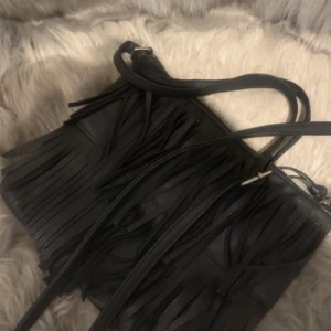 Svart axelväska med fransar i skinn - Cool svart axelväska i skinn med långa fransar på framsidan. Väskan har dragkedja upptill och en smal axelrem, perfekt för dig som vill ha en edgy och trendig accessoar. Enkel design men fransarna ger den en unik look.