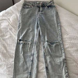 Ljusa ripped jeans från NA-KD - Säljer ett par ljusblå jeans från NA-KD med slitna hål över knäna. Jeansen har klassisk femficksmodell, raka ben och hög midja. Materialet är denim i bomull och de har en cool, avslappnad vibe med sina slitningar.