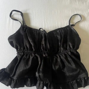 Svart volangig linnetopp med knyt - Supersöt svart linnetopp med smala axelband, volangkant nertill och knytning framtill. Toppen har en lätt glansig yta och resår i midjan för en snygg passform. 