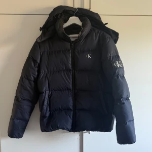 Marinblå pufferjacka från Calvin Klein - Säljer en marinblå vinterjacka från Calvin Klein  med huva och dragkedja. Jackan har broderad CK-logga på bröstet och en stor patch på ärmen. Perfekt för kalla dagar och riktigt snygg till streetwear-stilen. Väldigt bra skick. 