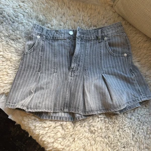 Grå randig jeanskjol från Zara XS - Säljer en grå, kort jeanskjol från Zara i storlek XS. Kjolen har tunna vita ränder, klassiska jeansfickor och hällor för bälte. Den är lätt utställd med snygga veck och stängs med dragkedja och knapp framtill.
