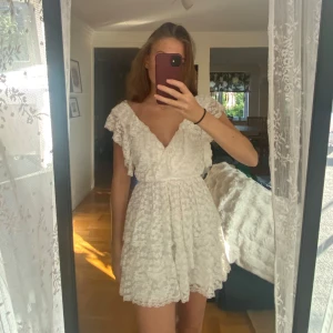 Vit spetsklänning med volang  - Supersöt vit kortklänning i spets med omlott och v-ringning. Klänningen har volangdetaljer, korta ärmar och knyt i ryggen. Perfekt på studenten eller vad som. Super bra skick såg inga tecken på användning 
