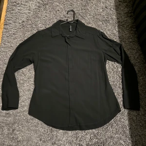 Svart skjorta från H&M med knappar - Klassisk svart skjorta från H&M med dold knäppning framtill och lång ärm. Skjortan har en rak passform, klassisk krage och är tillverkad i ett mjukt, lätt material som känns skönt mot huden. Perfekt för en clean och stilren look.