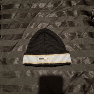Svart ribbad mössa från Reima - Svart ribbstickad mössa från Reima med uppvikt kant i ljusgrått och beige. Märkeslogga framtill på kanten. Perfekt för kyliga dagar och enkel att matcha med olika outfits.
