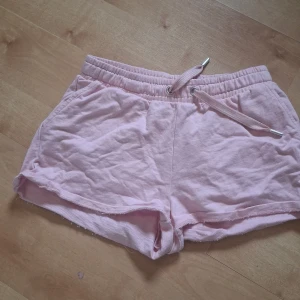 Rosa shorts med rosett från Lindex - Söta ljusrosa shorts från Lindex med snörning i midjan och två bakfickor dekorerade med broderade rosetter. Mjukt bomullsmaterial och avslappnad passform, perfekta för varma sommardagar.