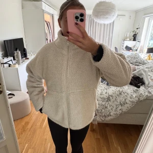 Beige teddyjacka med dragkedja - Mysig beige teddyjacka med hög krage och hel dragkedja framtill. Jackan har en loose passform och är tillverkad i fluffigt fleece-material.