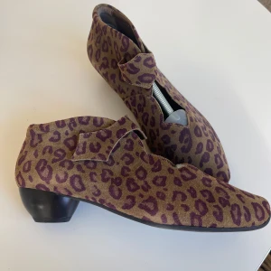 Leopardmönstrade skor från Think i skinn och mocka - Unika skor från Think i skinn och mocka med leopardmönster i lila och brunt. Skorna har låg klack, spetsig tå och dekorativ knäppning framtill. Gummisula.  Perfekta för dig som vill sticka ut med ett djurmönstrat statement. Tillverkade i Italien.