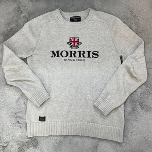 Grå stickad tröja från Morris - Snygg ljusgrå stickad tröja från Morris med broderad logga och brittisk flagga framtill. Tröjan har rund hals, långa ärmar och ribbade muddar vid ärmslut och nederkant. Perfekt för dig som gillar klassisk och stilren look.
