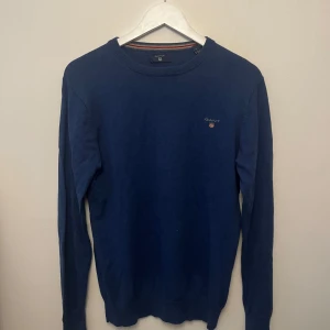 Mörkblå bomullströja från GANT - Snygg mörkblå tröja från GANT i bomull med rund halsringning och diskret logga på bröstet. Klassisk modell med ribbade muddar och lång ärm, perfekt för en stilren look.