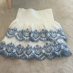 Vit kjol med blå broderi från Lindex - Supersöt vit kjol från Lindex med bred smock i midjan och två lager volanger med blå broderade detaljer. Kjolen har en luftig och flowy känsla, perfekt för dig som gillar romantisk stil och snygga broderier.