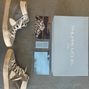 Philippe Model grå höga sneakers - Snygga höga sneakers från Philippe Model i grå mocka med vita detaljer och snörning. Skorna har en klassisk siluett med rund tå och platt sula. Kommer med originalkartong och märkets emblem på sidan. Perfekta för dig som gillar stilrena och exklusiva sneakers. Edast skorna medföljer