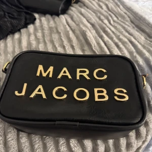 Svart Marc Jacobs handväska i skinn - Pris kan diskuteras Snygg svart handväska från Marc Jacobs i skinn med stora guldiga bokstäver på framsidan. Väskan har en rektangulär form och detaljer i guld. Perfekt för dig som vill ha en stilren och trendig accessoar.