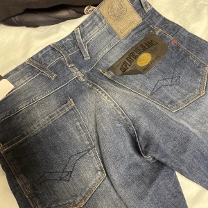 Replay Anbass blå jeans W28 L32 - Säljer ett par Replay Anbass jeans i klassisk blå tvätt. Jeansen har raka bakfickor med snygga sömmar, Replay-logga på läderpatch och detaljerad etikett. Tillverkade i slitstarkt bomullsmaterial och passar perfekt till en avslappnad streetstil.