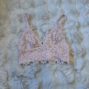 Ljusrosa spetsig linnetopp från BikBok - Supersöt ljusrosa linnetopp från BikBok med spetsdetaljer och justerbara axelband. Toppen har blommigt spetsmönster framtill och slät baksida i glansigt material. Perfekt för att ge outfiten en romantisk touch.