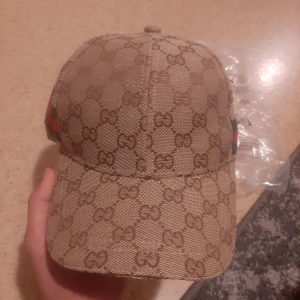 keps med GG-mönster och rödgrön detalj - Snygg keps från  i beige med klassiskt GG-monogram över hela kepsen. Kepsen har en böjd skärm och en ikonisk rödgrön rand på sidan. Tillverkad i ullblandning med merinoull och kashmir. Perfekt accessoar för att lyfta din stil.