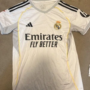 Real Madrid fotbollströja Adidas M - Snygg Real Madrid fotbollströja från Adidas i vitt med svarta detaljer och gula linjer. Tröjan har klubbmärke och sponsortryck på bröstet, klassiska Adidas-ränder på axlarna och är tillverkad i lätt, ventilerande material. Perfekt för dig som älskar fotboll och vill bära ditt lag.
