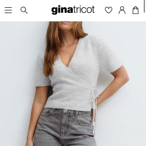 Ljusgrå omlottopp från Gina Tricot - Supermjuk ljusgrå omlottopp med kort ärm och knyt i sidan. Toppen har v-ringning och en lite fluffig, stickad känsla som gör den extra mysig. Perfekt att styla med jeans eller kjol för en chill look.