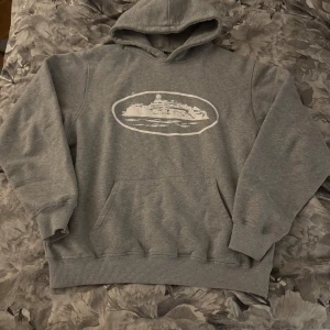 Grå corteiz hoodie - Säljer en grå corteiz hoodie med vit tryckt illustration av Alcatraz på bröstet. Tröjan har huva, känguruficka och ribbade muddar. Perfekt för dig som gillar streetwear och vill ha något unikt i garderoben.