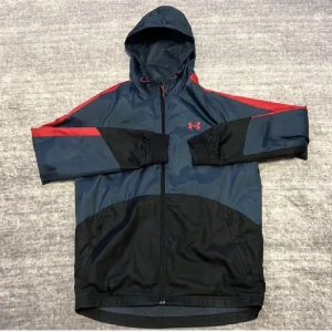 Under Armour vindjacka blå/röd S - Sportig vindjacka från Under Armour i mörkblått och svart med röda detaljer och logga. Jackan har huva med dragsko, hel dragkedja och meshfoder för extra komfort. Perfekt för aktiva dagar och snygg streetstil.