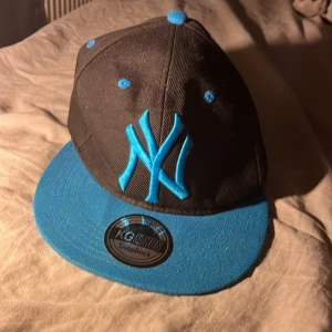 Svart och blå New York Yankees keps - Säljer en snapback-keps från New York Yankees med svart krona och blå skärm. Kepsen har blå broderade detaljer, inklusive NY-loggan fram och MLB-loggan bak. Justerbar storlek med blå plastrem. Perfekt för dig som vill ha sportig streetstyle.