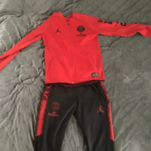 PSG x Jordan röd dress - Röd ziptröja från PSG x Jordan med svarta detaljer och loggor på bröstet. Långärmad modell med dragkedja hela vägen och fickor på sidorna. Perfekt för dig som älskar fotboll och streetstyle.