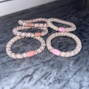 Rosa glittriga armband med pärldetalj - Fem stycken elastiska armband i ljusrosa och guldglittrande flätad design, med en oval pärldetalj i rosa plast. Perfekta för att ge en söt och trendig touch till din outfit.