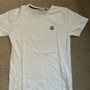 Vit t-shirt från Moncler, storlek S - Vit t-shirt från Moncler i mjuk bomull med klassisk rund hals och korta ärmar. Diskret Moncler-logga broderad på bröstet. Enkel och clean design som passar perfekt till jeans eller shorts.