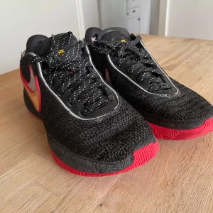 Svarta Nike leBron 20 skor med röd och guld swoosh - Säljer ett par svarta Nike leBron 20 med stickad ovandel, röda detaljer och en metallic guld swoosh. Skorna har svart sula med röd undersida och svarta snören med vita prickar. Perfekta för dig som vill sticka ut med en unik och sportig look.