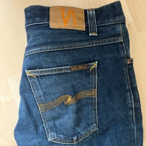 Nudie Jeans Steady Eddie II Dry Platino - Mörkblå jeans från Nudie Jeans, modell Steady Eddie II Dry Platino. Klassisk femficksdesign med orange kontrastsömmar och Nudie-logga på bakfickan. Tillverkade i 100% bomull för en autentisk jeanskänsla och rak passform.