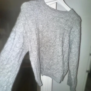 Grå stickad tröja från H&M Divided - Mysig grå stickad tröja från H&M Divided med kabelstickat mönster. Tröjan har rund halsringning, långa ärmar och ribbade muddar. Perfekt för lager-på-lager under kyliga dagar.