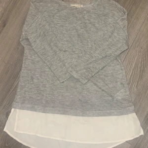 Långärmad tröja från 157 Angelology - Grå långärmad tröja från 157 Angelology med vit chiffongliknande nederdel som ger en lager-på-lager-look. Tröjan har rund halsringning och är i mjuk bomullsblandning. 