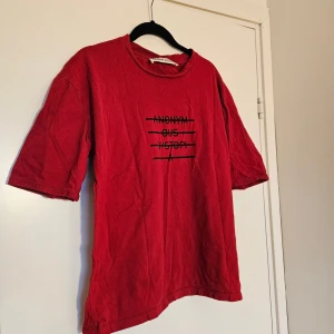 Röd t-shirt från Adnym Atelier - Cool röd t-shirt från Adnym Atelier med svart tryck på bröstet där det står 'ANONYM OUS DYSTOPI' med överstrykningar. Oversized passform och klassisk rund hals. 