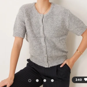 Grå stickad kofta med pärlknappar - Mysig grå stickad kofta med korta ärmar och rund hals. Koftan har pärlknappar framtill och är tillverkad i ett fluffigt, mjukt material som ger en chill och trendig vibe. Perfekt att slänga över en topp för extra stilpoäng.