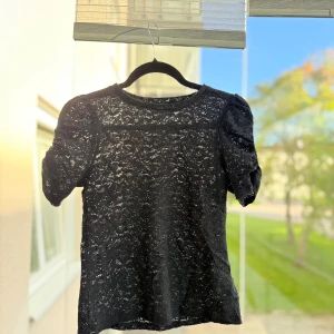 Svart spetsblus med puffärm från Kaphal - Säljer en svart blus i genombruten spets med korta puffärmar. Från Kappahl.  Toppen har rund halsringning och ett vackert blommigt spetsmönster. 