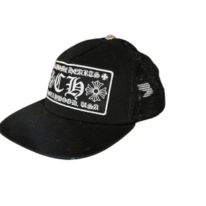 Chrome Heart keps  - Svart truckerkeps från Chrome Hearts med vit broderad logga och blommotiv framtill. Kepsen har mesh på sidorna och bak för extra ventilation samt en böjd skärm. Snygg detalj med metallknopp på toppen.