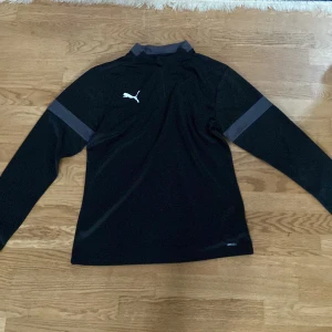 Svart långärmad träningströja  - Svart långärmad träningströja från Puma med halv dragkedja framtill och gråa detaljer på ärmarna. Tillverkad i ett lätt och funktionellt polyestermaterial som andas. Perfekt för träning eller sportiga dagar.