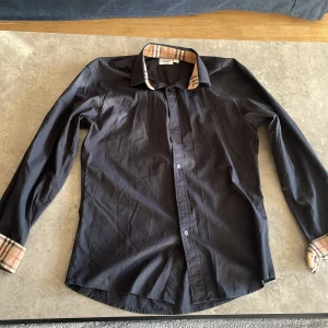 Svart Burberry skjorta med rutiga detaljer - Svart skjorta från Burberry i bomull med klassiska rutiga detaljer på krage och ärmslut. Skjortan har långa ärmar, knappar framtill och broderad B-logga på bröstet. Perfekt för dig som gillar stilrena plagg med en twist.