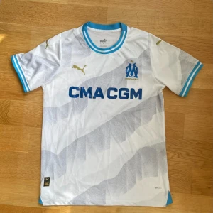 Marseille Puma fotbollströja vit/blå - Säljer en officiell Olympique de Marseille fotbollströja från Puma. Tröjan är vit med ljusgråa mönster, blå detaljer vid krage och ärmslut samt guldiga Puma-loggor. Materialet är lätt och andas, perfekt för match eller träning. Tryckt sponsorlogga på bröstet.