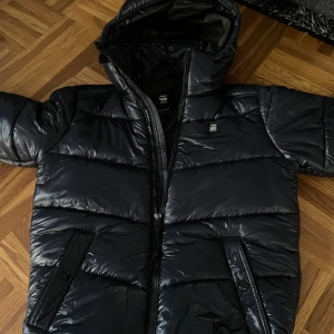 Svart pufferjacka från G-Star RAW - Snygg svart pufferjacka från G-Star RAW med glansig finish och huva. Jackan har dragkedja framtill, två fickor med knappdetaljer och diskret logga på bröstet. Perfekt för kalla dagar när du vill hålla stilen.