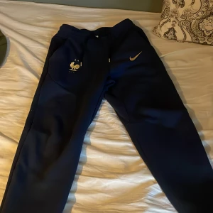 Blå Nike träningsbyxor Frankrike S - Mörkblå träningsbyxor från Nike med Frankrikes landslagslogga och guldfärgad swoosh. Byxorna har dragkedja vid fickan och är tillverkade i ett mjukt syntetmaterial. Perfekta för dig som gillar sportig stil och vill representera Frankrike.