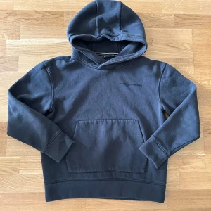 Mörkblå hoodie från Sail Racing - Mörkblå hoodie från Sail Racing med broderad logga på bröstet och en stor känguruficka framtill. Tröjan har huva och ribbade muddar vid ärmslut och nederkant. Perfekt för dig som gillar en stilren och sportig look.